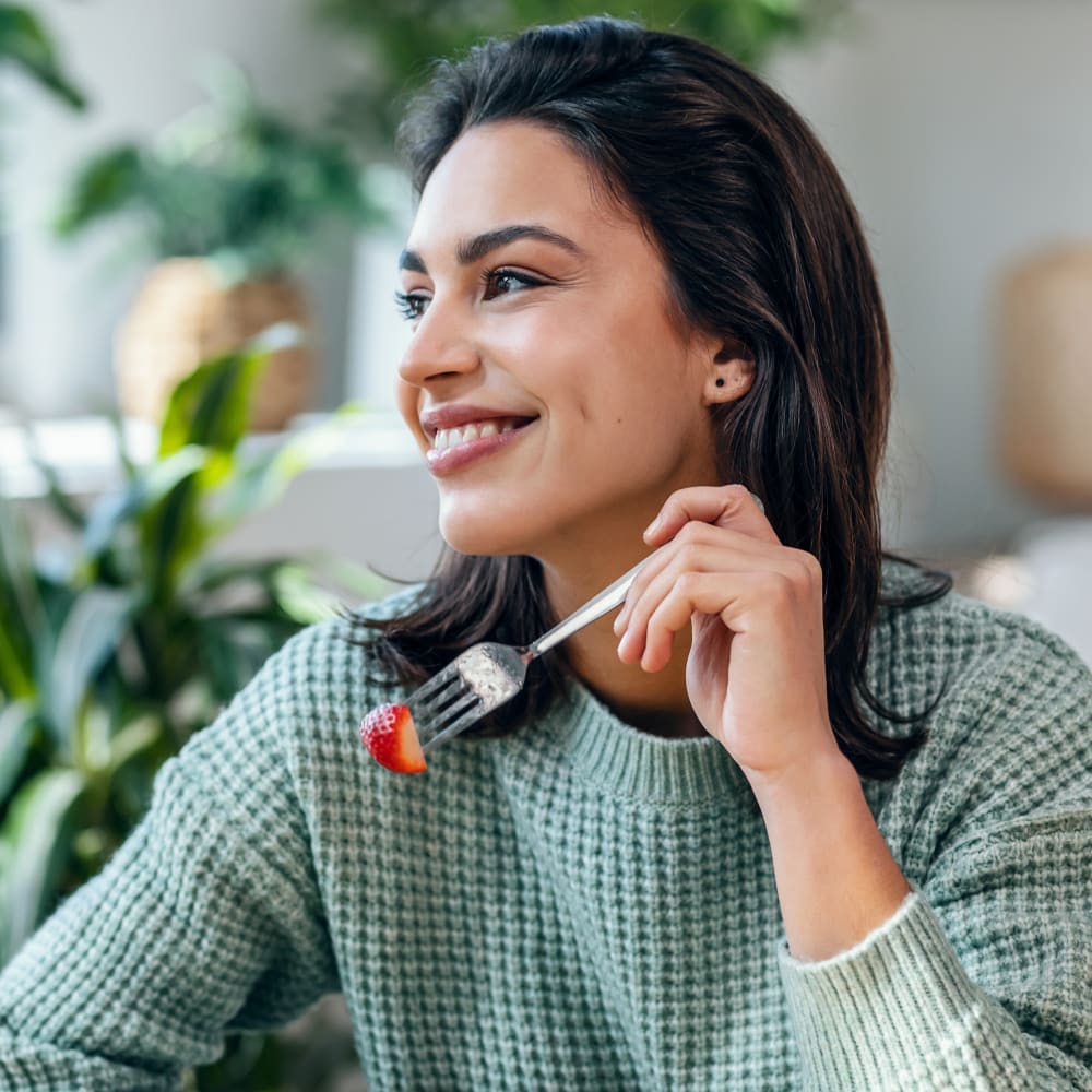 A woman wearing a green sweater looks to her right while holding up a strawberry on a fork. There are green, leafy plants in the background.
