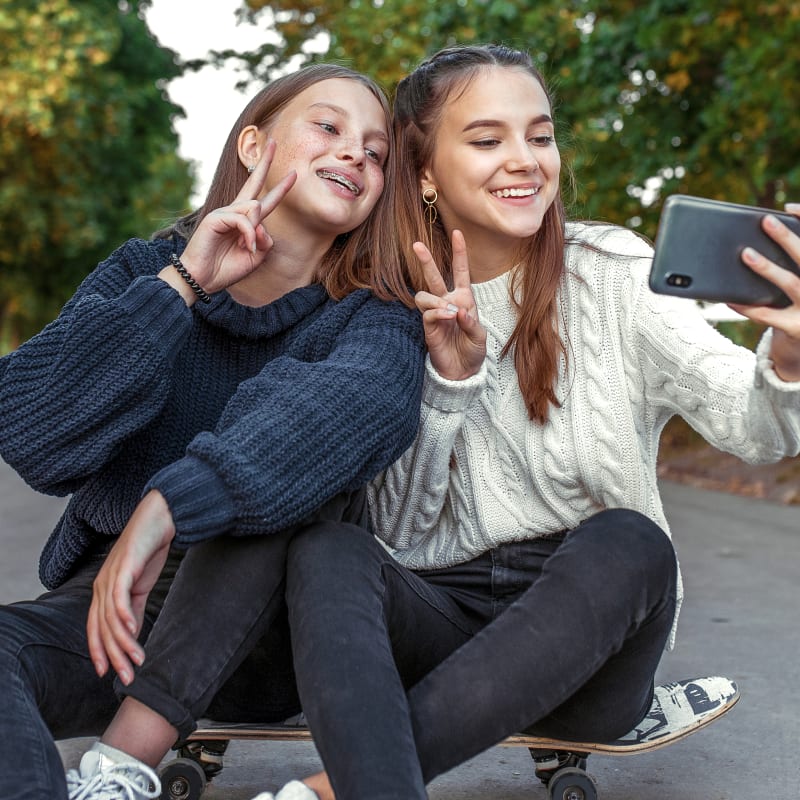 Two teenage girls wearing sweaters and black jeans are sitting on a skateboard. One girl has braces and they are both making a peace sign with their fingers while taking a selfie on a cell phone.