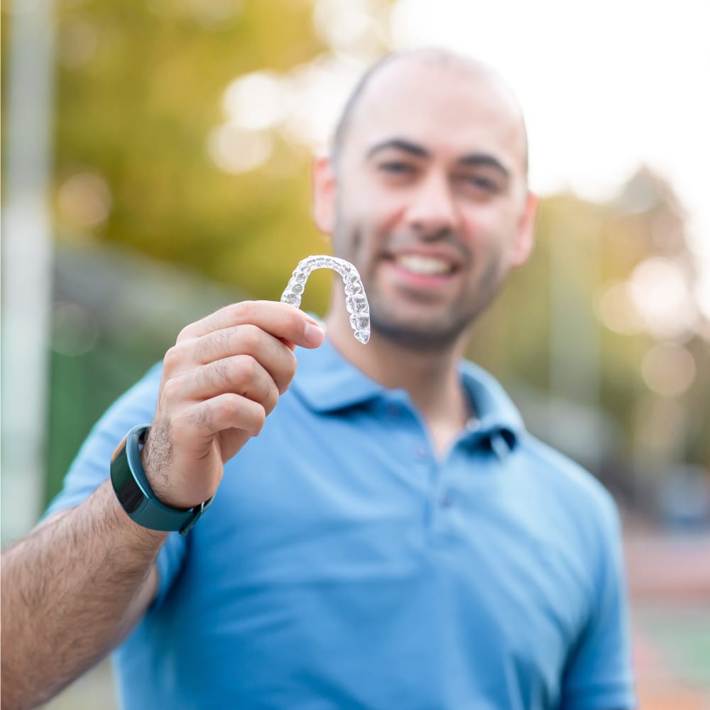 Man standing outside in front of trees wearing a light blue t-shirt and dark colored digital watch. He is lifting up his right hand holding a clear aligner tray.
