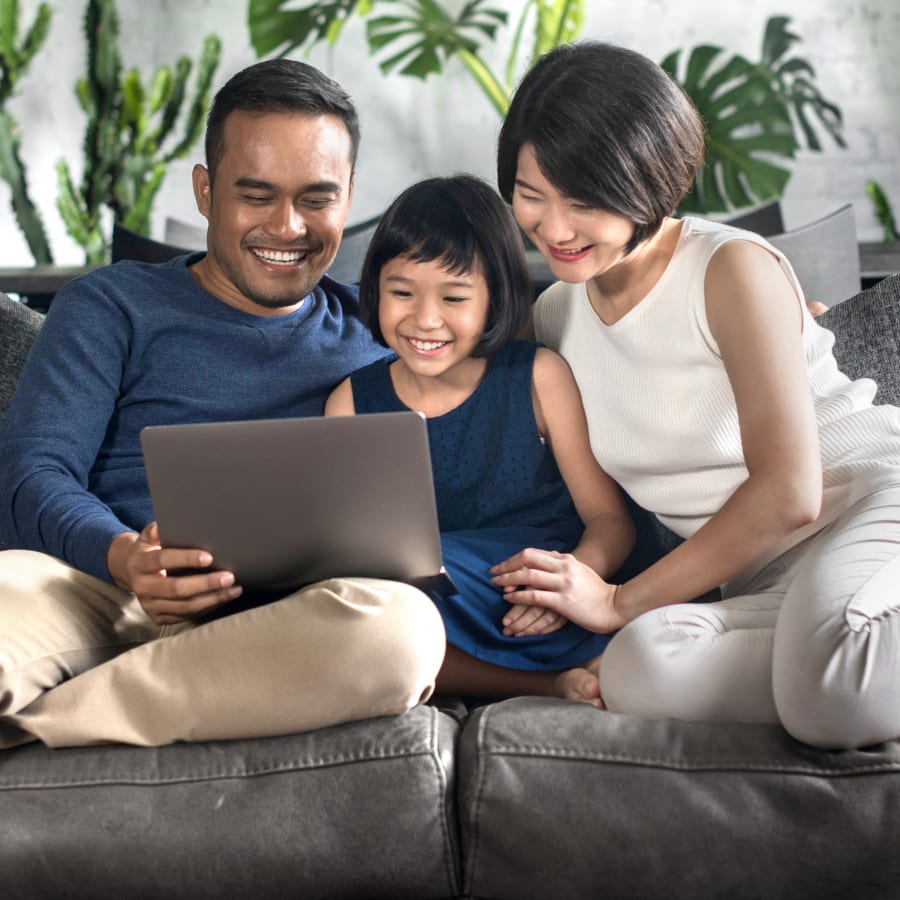 A family sitting outside with green trees in the background. The young boy is sitting on his fathers lap while the mother  sits to the left of them with the sister to the left of the mother.
